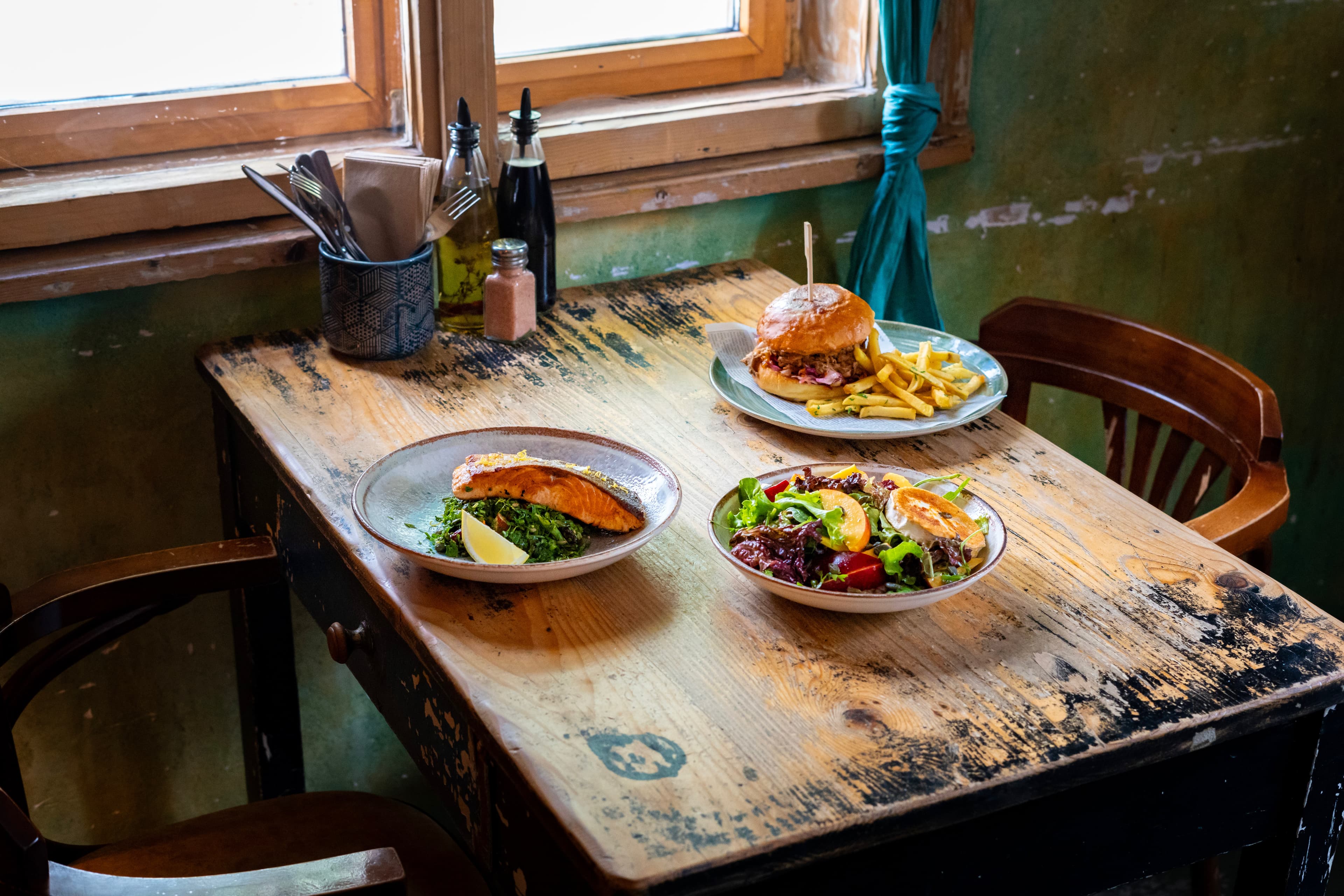 Three dishes on a wooden table by the window — salmon on greens, burger with fries and a summer salad