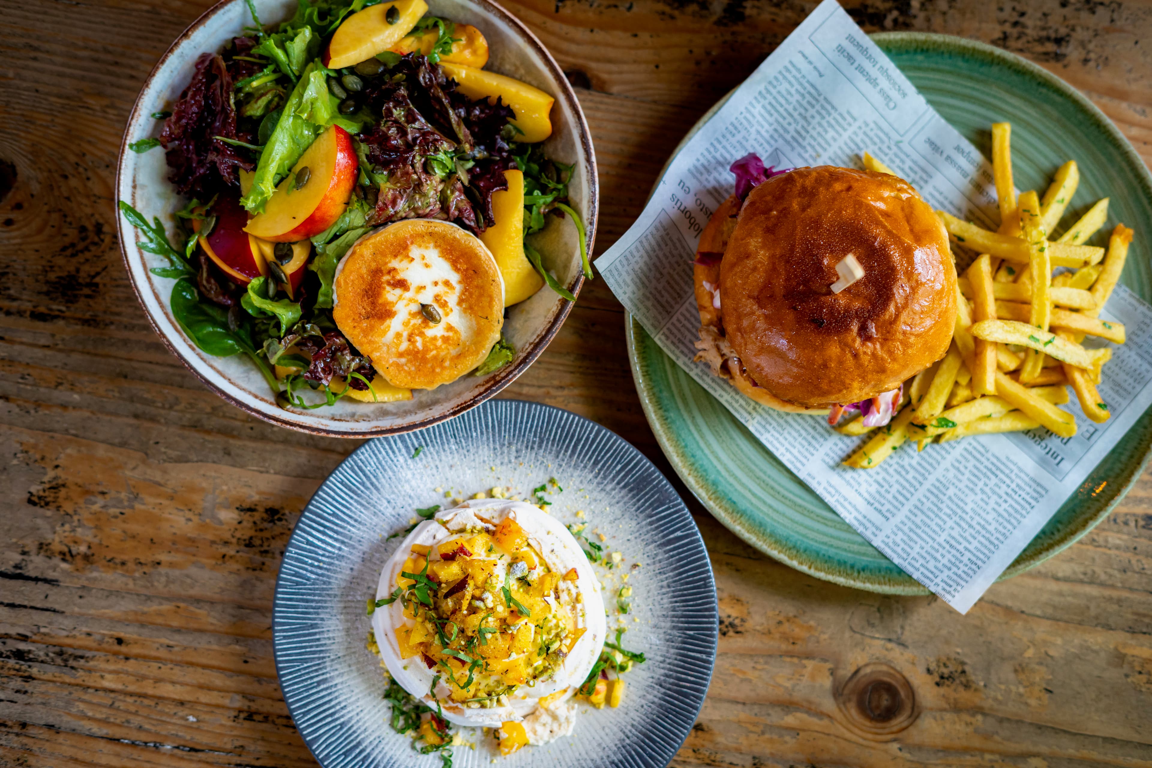 Overhead spread of mixed-leaf salad with goat cheese, pulled-pork burger on newspaper and a creamy mango dish