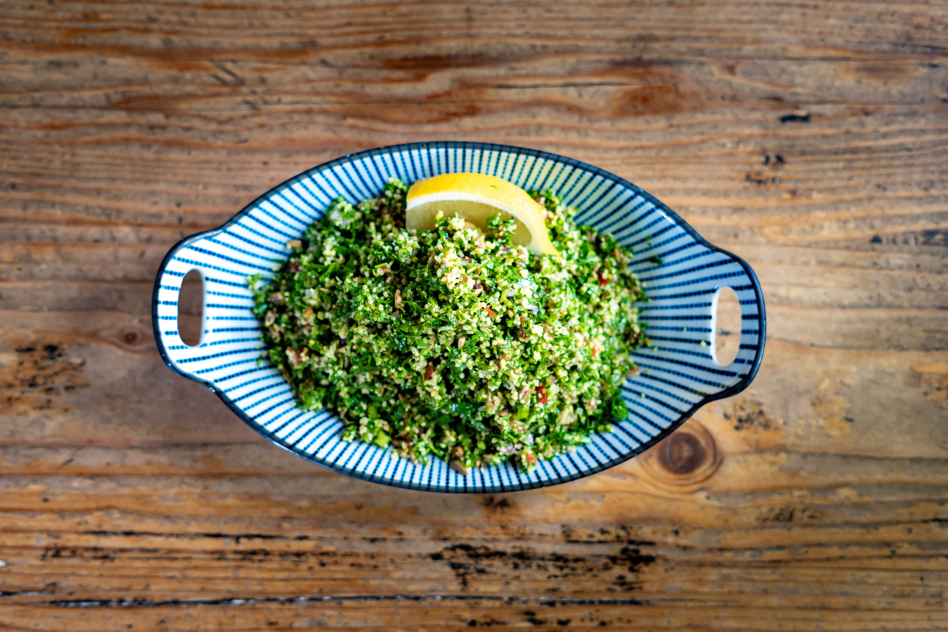 Overhead shot of herb tabbouleh salad in a blue-striped oval bowl with a lemon wedge