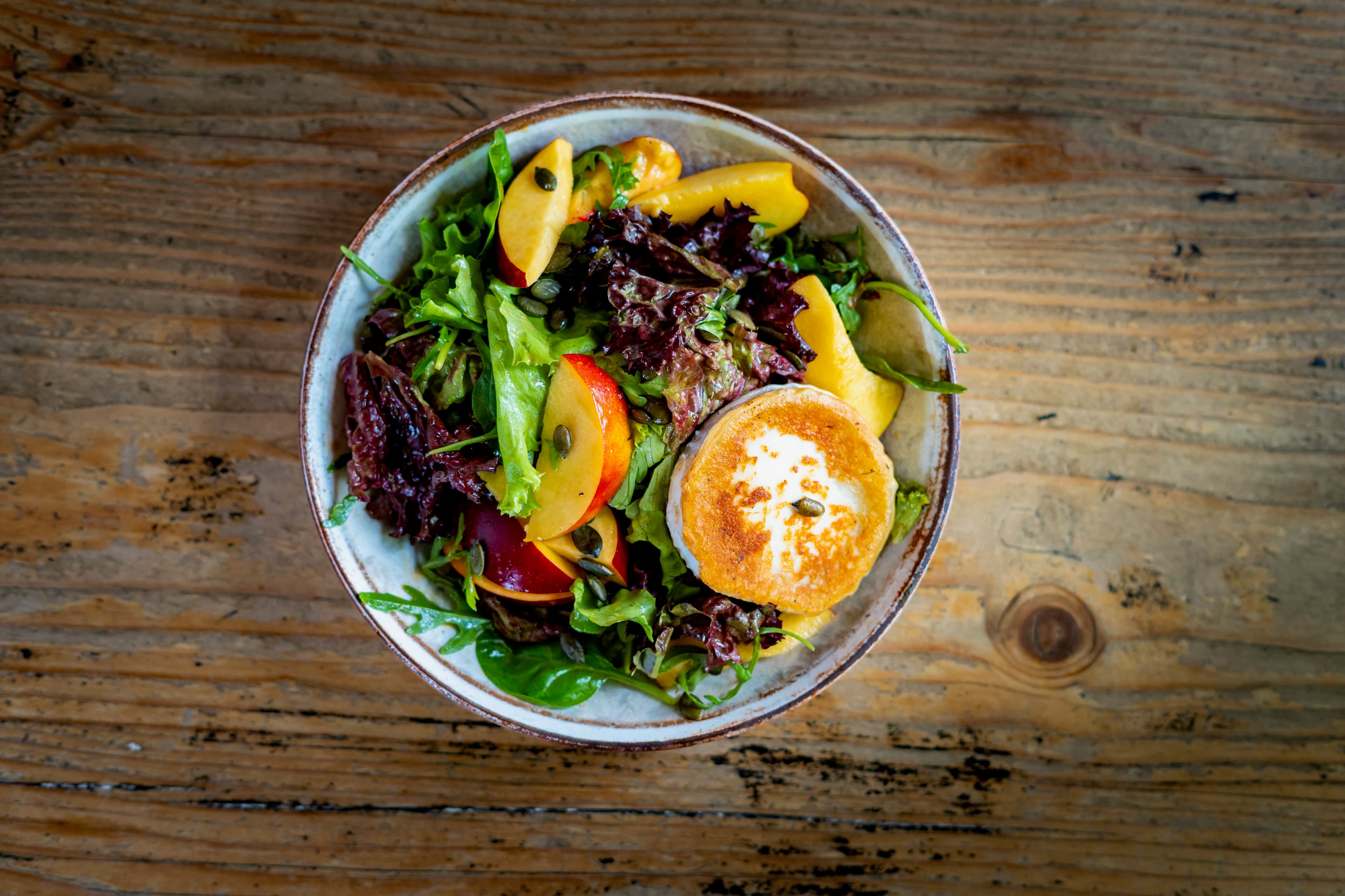 Overhead bowl of mixed leaves with peach slices and a warm goat cheese medallion
