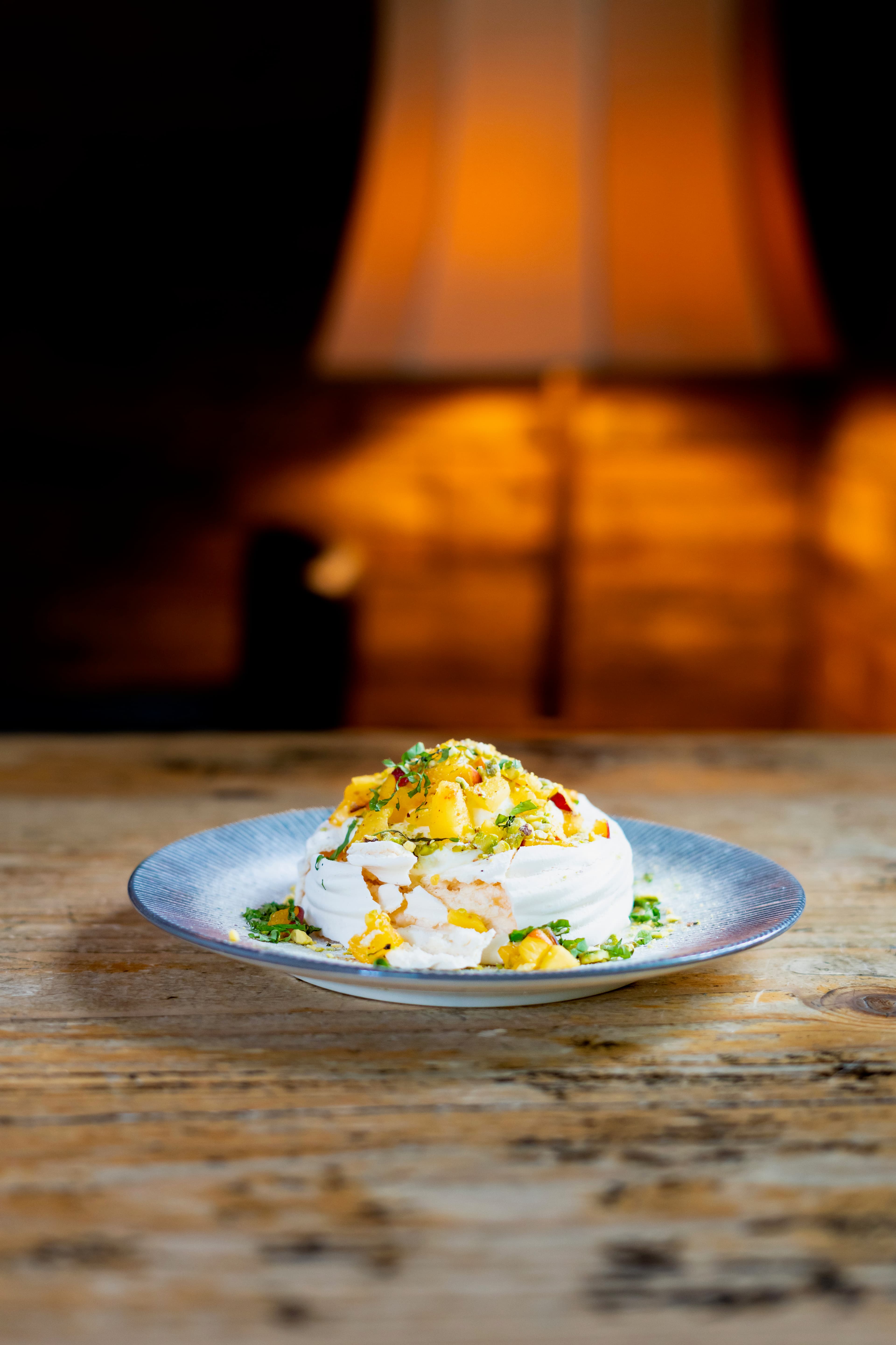 Pavlova with mango and herb topping on a blue plate, warm lamp glow in the background