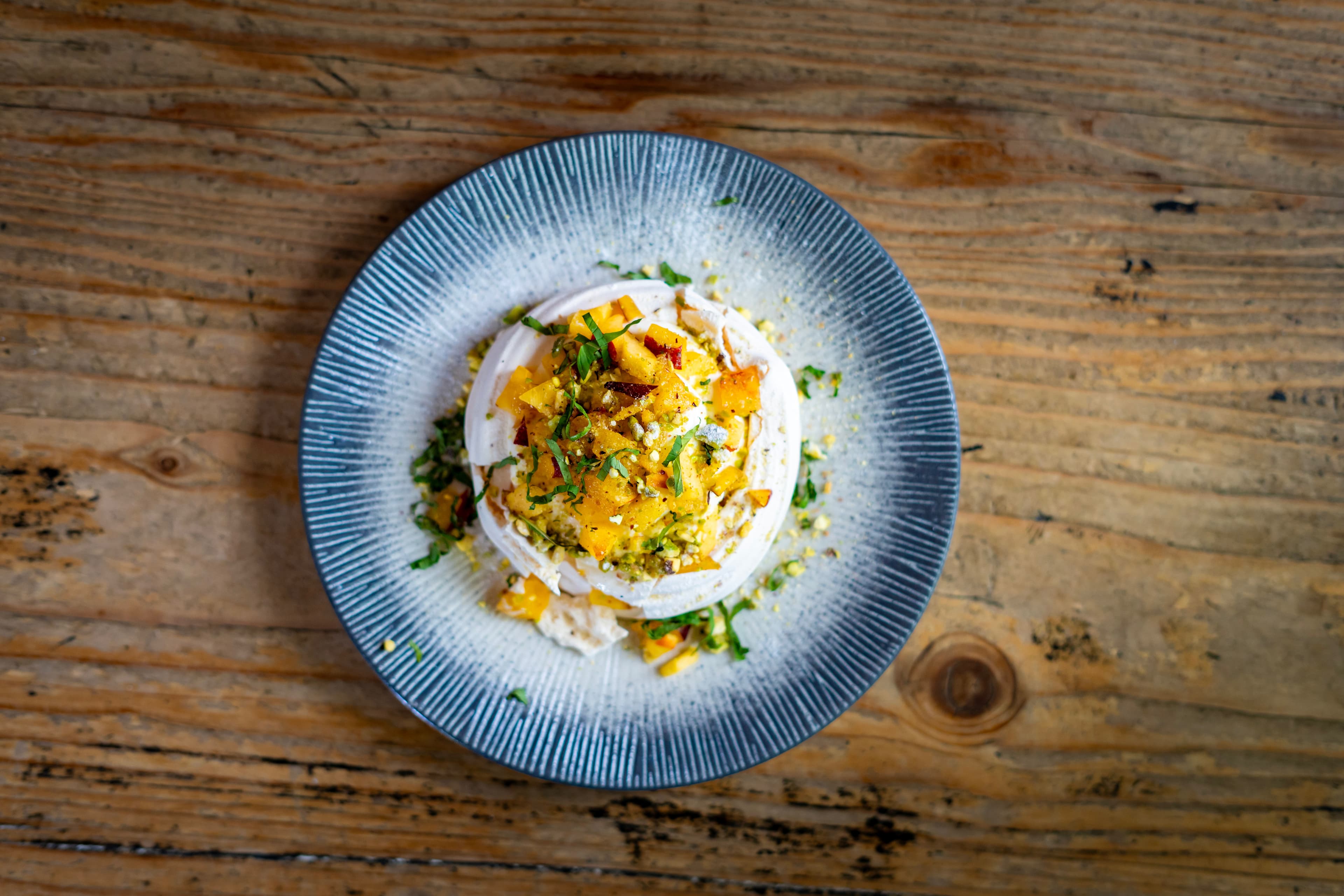 Overhead pavlova with mango and chopped herbs on a blue ribbed plate, wooden table